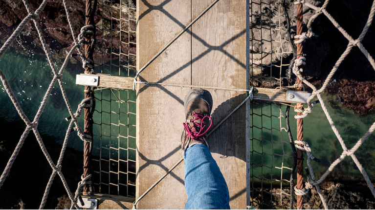 Overhead view of a visitor wearing brown hiking boots with pink laces and blue jeans, walking along Carrick-a-Rede Rope Bridge, with the crystal blue waters below